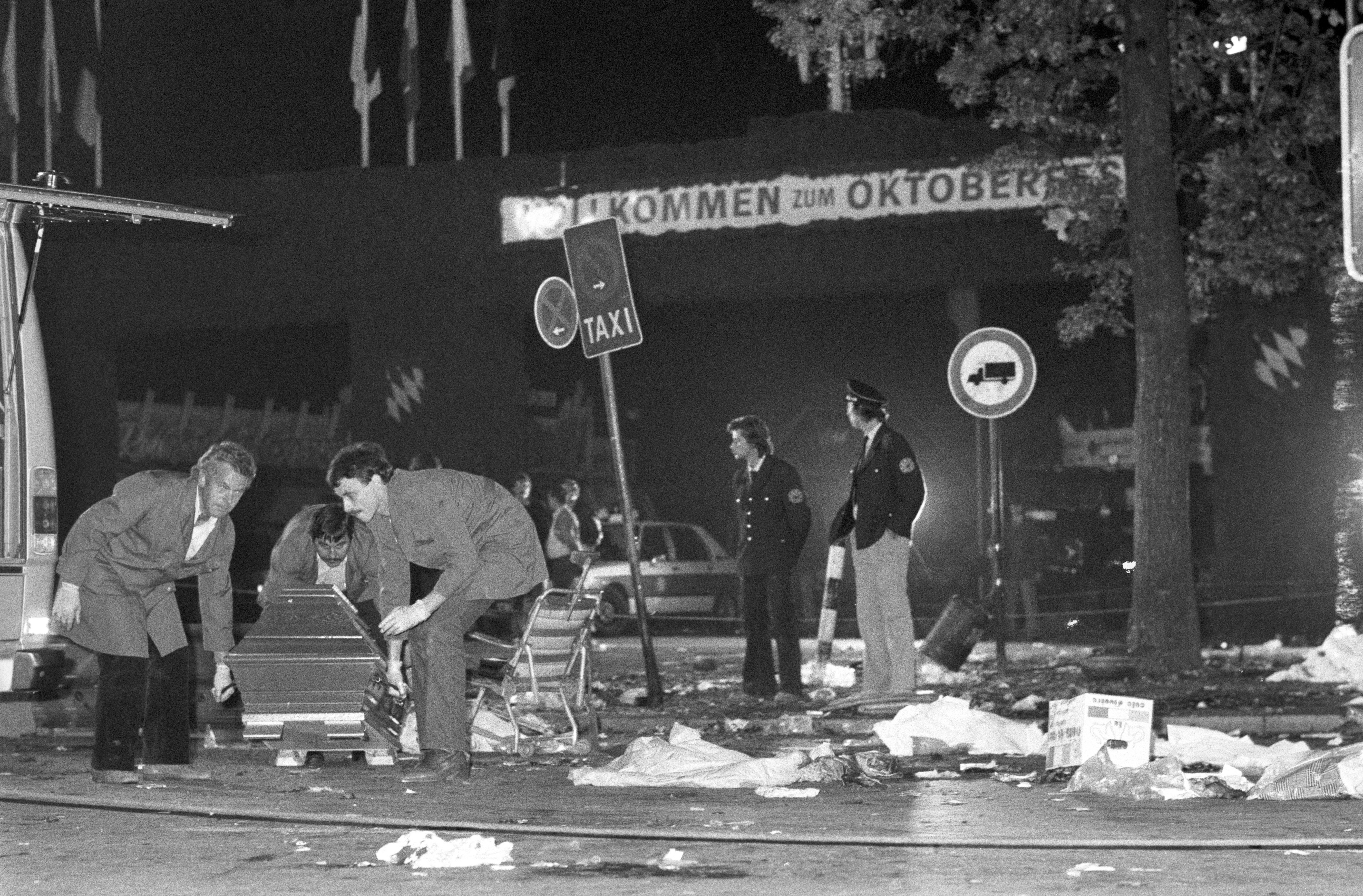 


                 Verwüsteter Haupteingang des Oktoberfests mit zwei Polizeibeamten und einem Polizeiauto. Links im Bild heben drei Männer einen Sarg in ein Fahrzeug.